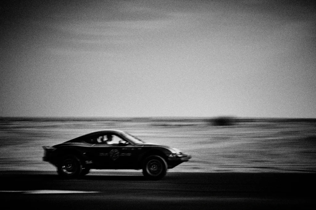 Grainy black-and-white panning photograph of an off-road Porsche Cayman driving along a desert road.