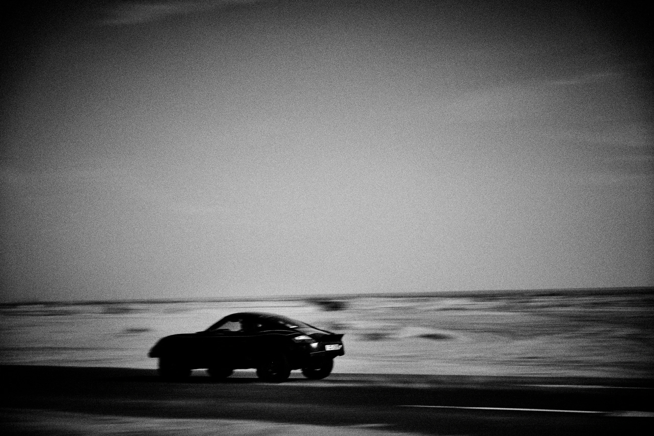 Grainy black-and-white panning photograph of an off-road Porsche Cayman driving along a desert road.