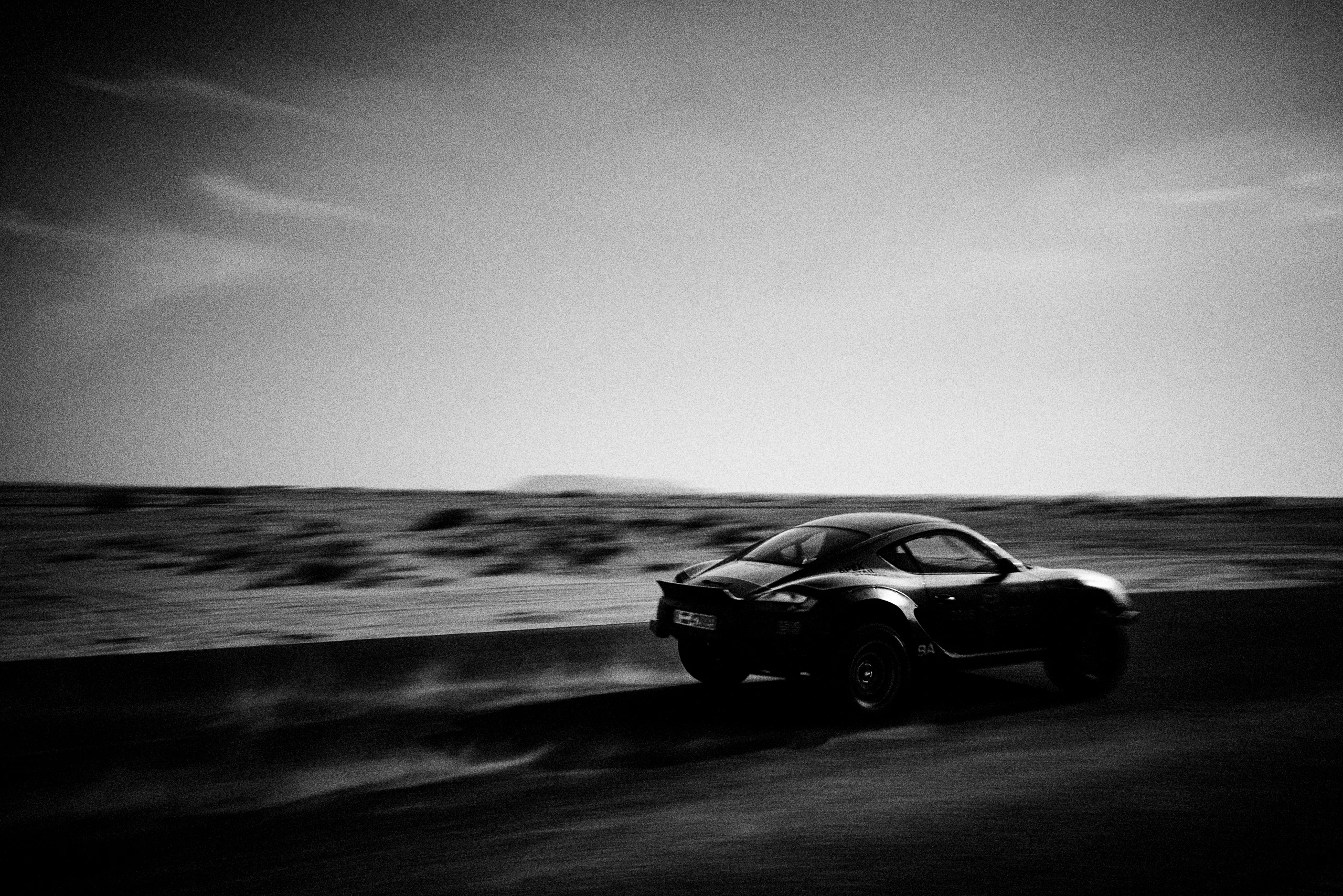 Grainy black-and-white panning photograph of an off-road Porsche Cayman driving along a desert road.