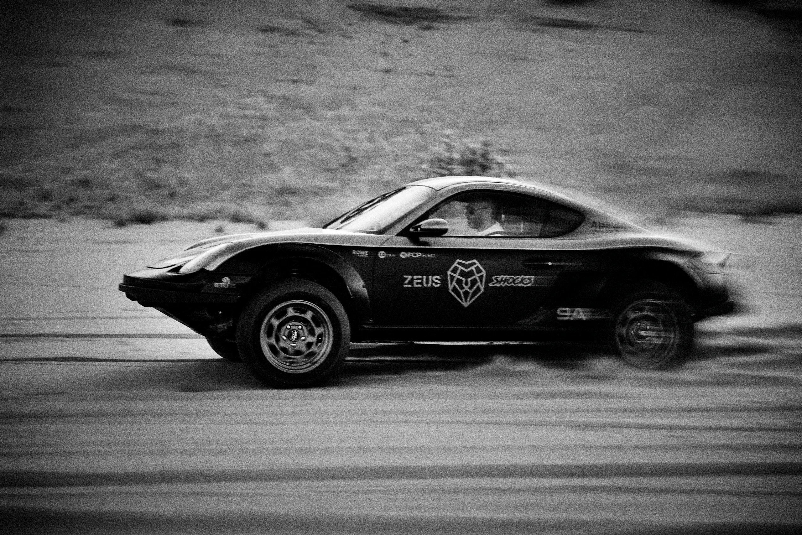Grainy black-and-white panning photograph of an off-road Porsche Cayman driving along a desert road.