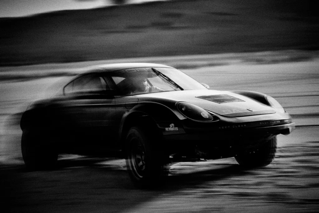 Grainy black-and-white panning photograph of an off-road Porsche Cayman driving along a desert road.