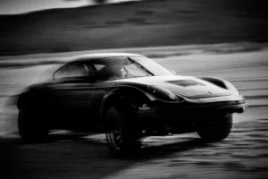 Grainy black-and-white panning photograph of an off-road Porsche Cayman driving along a desert road.