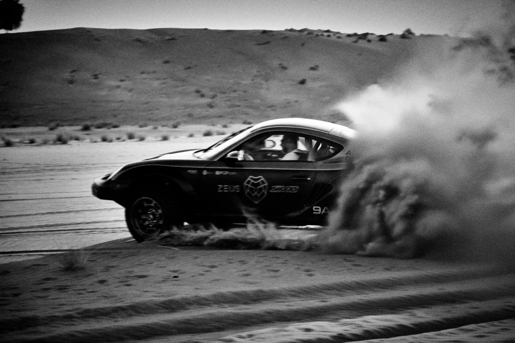 Grainy black-and-white panning photograph of an off-road Porsche Cayman driving along a desert road.