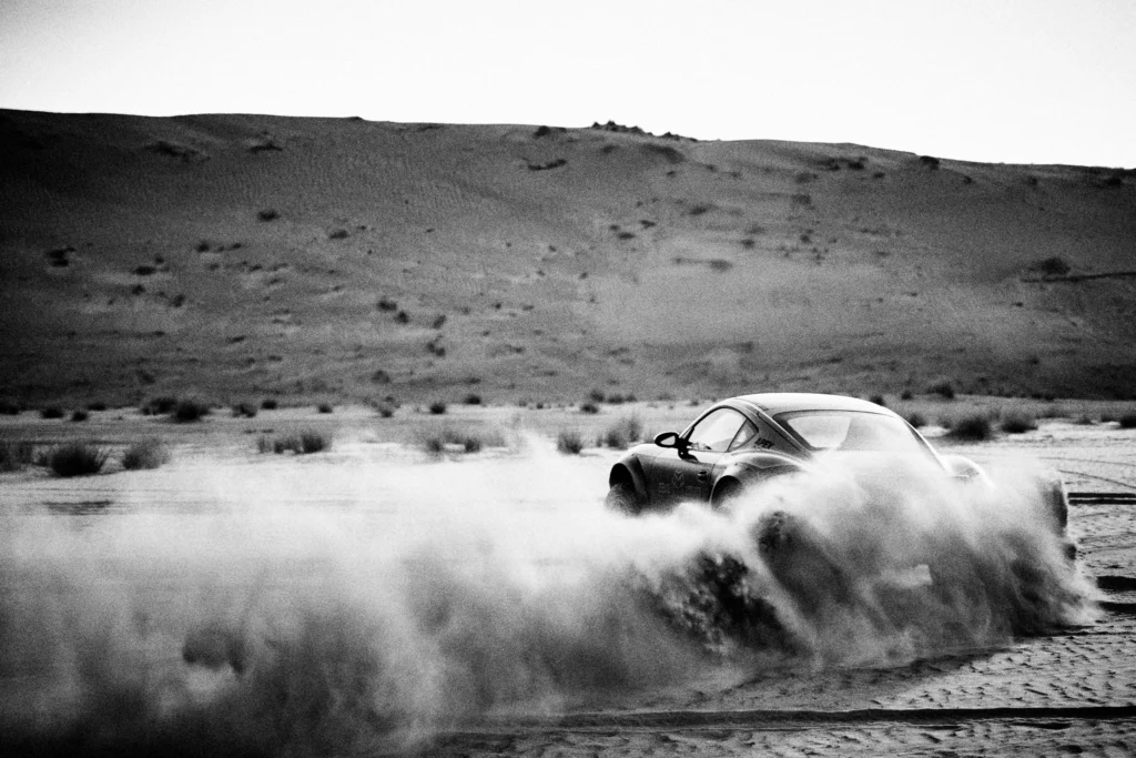 Grainy black-and-white panning photograph of an off-road Porsche Cayman driving along a desert road.