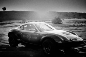 Grainy black-and-white panning photograph of an off-road Porsche Cayman driving along a desert road.