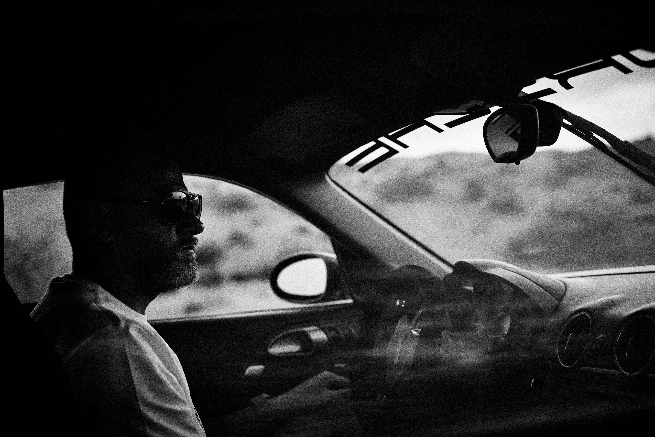 Grainy black-and-white panning photograph of an off-road Porsche Cayman driving along a desert road.