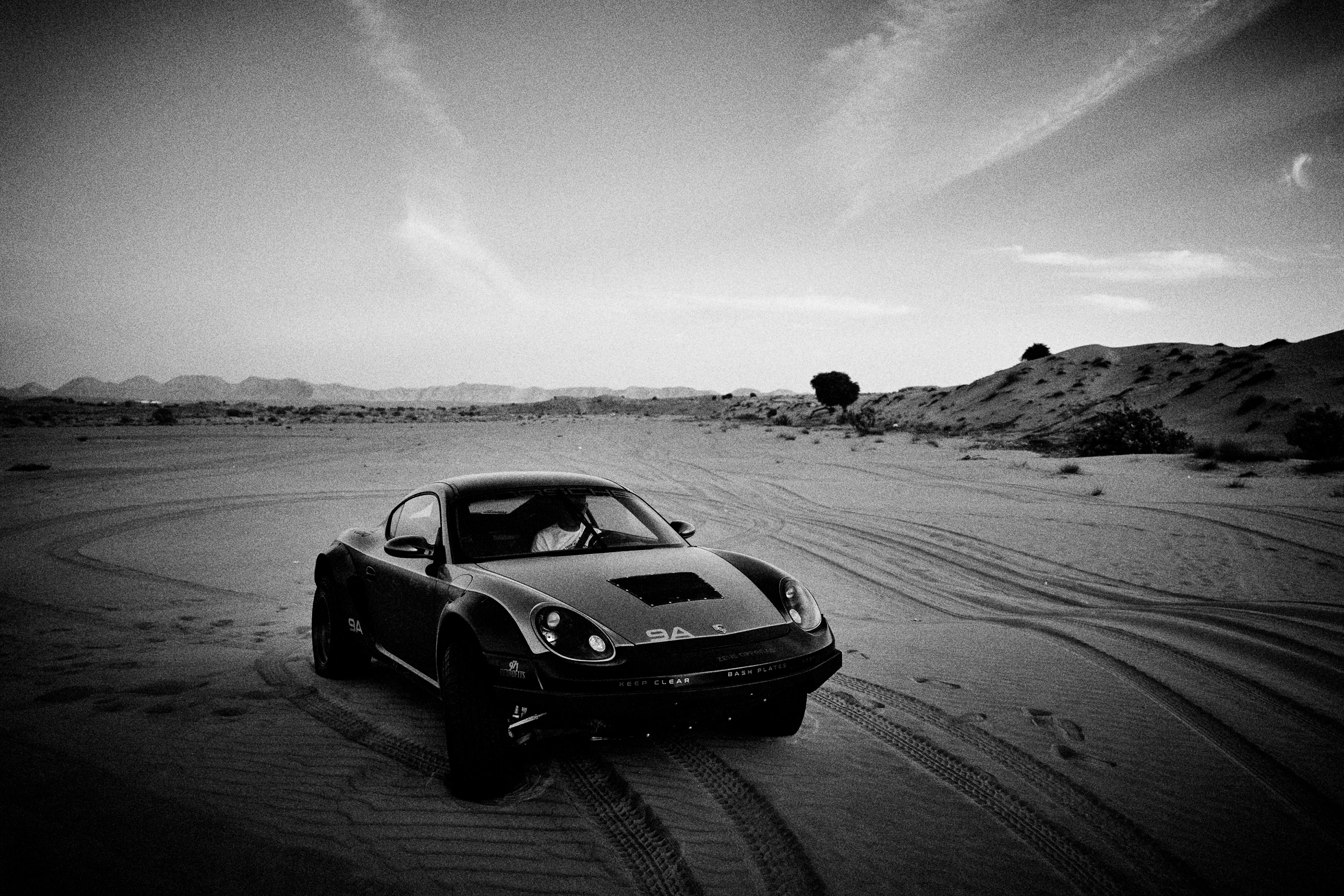 Grainy black-and-white panning photograph of an off-road Porsche Cayman driving along a desert road.