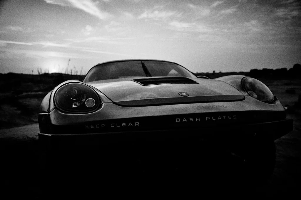 Grainy black-and-white panning photograph of an off-road Porsche Cayman driving along a desert road.