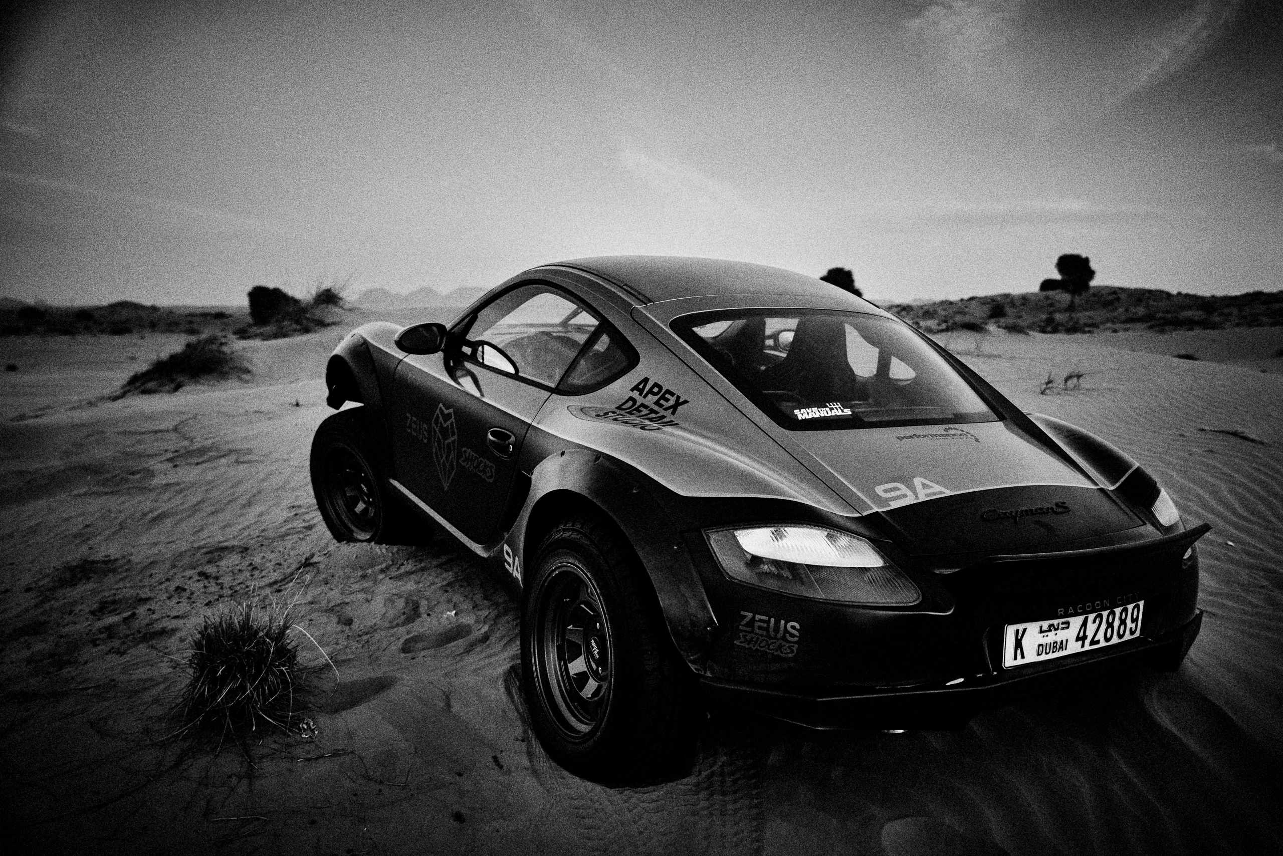 Grainy black-and-white panning photograph of an off-road Porsche Cayman driving along a desert road.