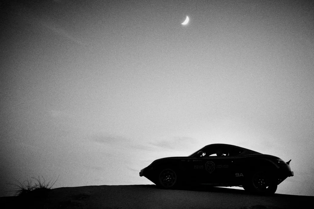 Grainy black-and-white panning photograph of an off-road Porsche Cayman driving along a desert road.