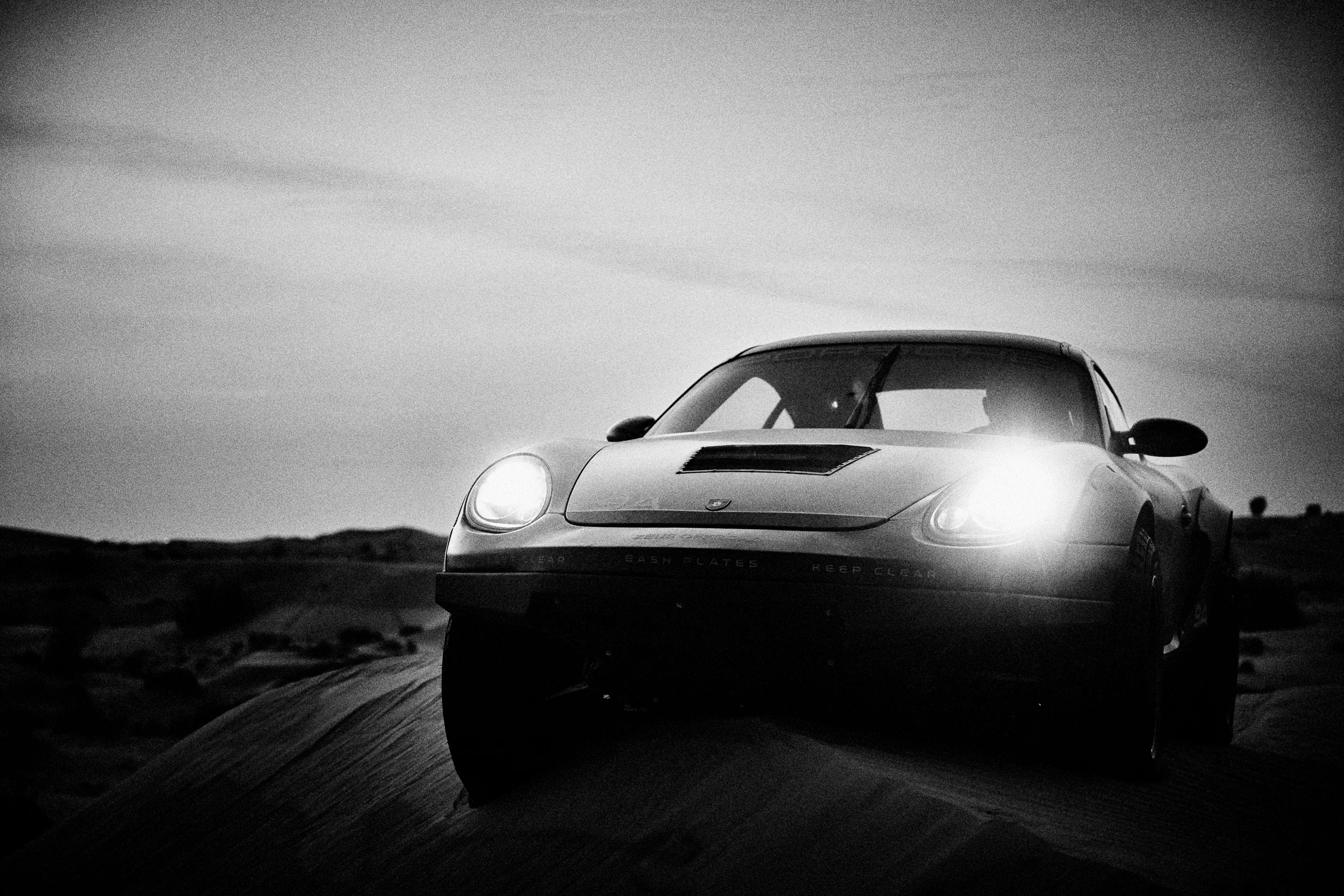 Grainy black-and-white panning photograph of an off-road Porsche Cayman driving along a desert road.