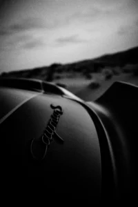 Grainy black-and-white panning photograph of an off-road Porsche Cayman driving along a desert road.