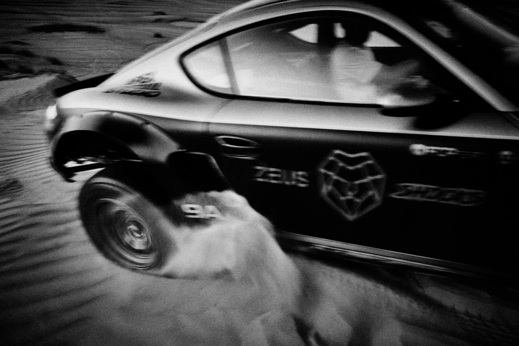 Grainy black-and-white panning photograph of an off-road Porsche Cayman driving along a desert road.