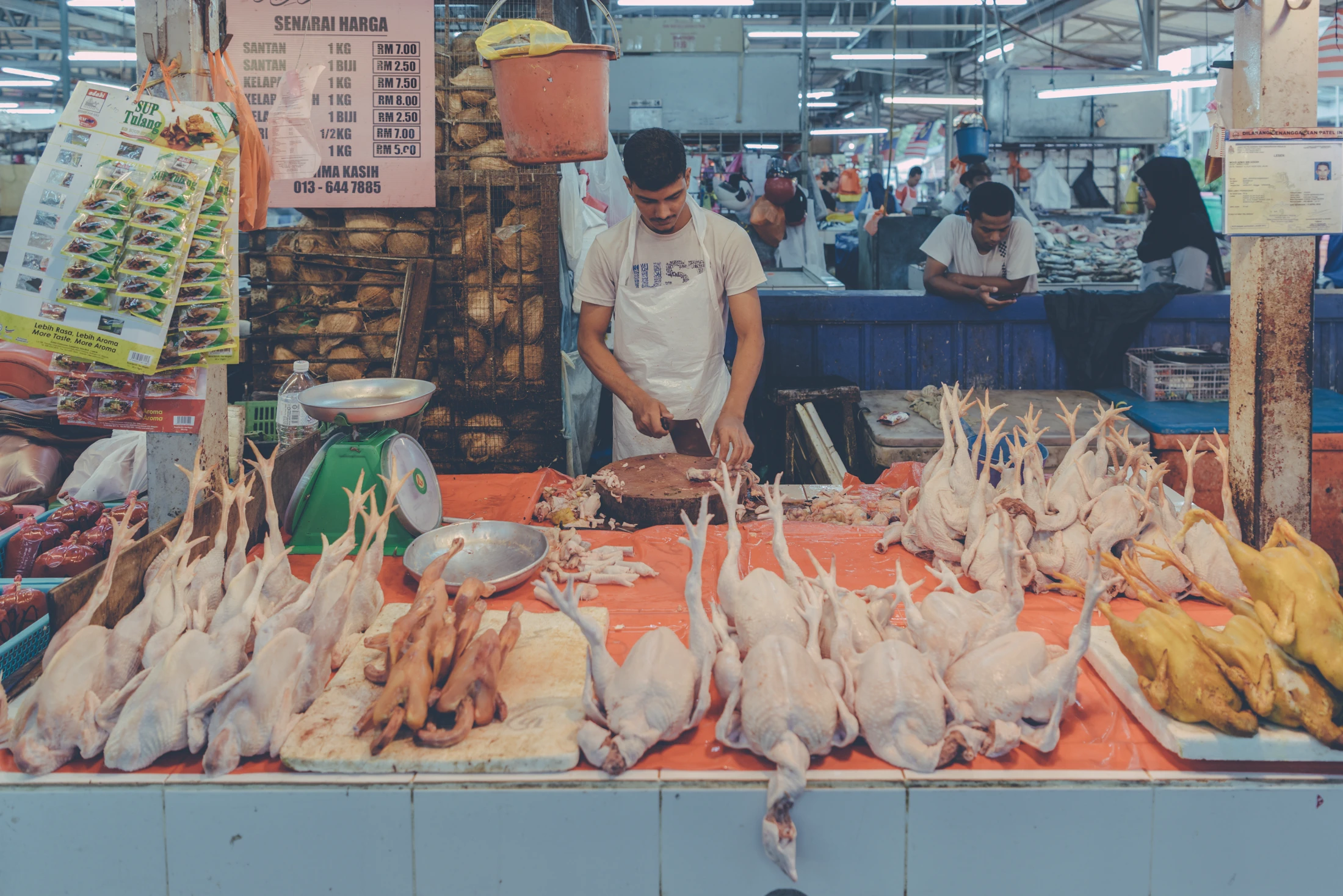 Poultry vendor behind a market counter covered with prepared whole chickens in Malaysia.