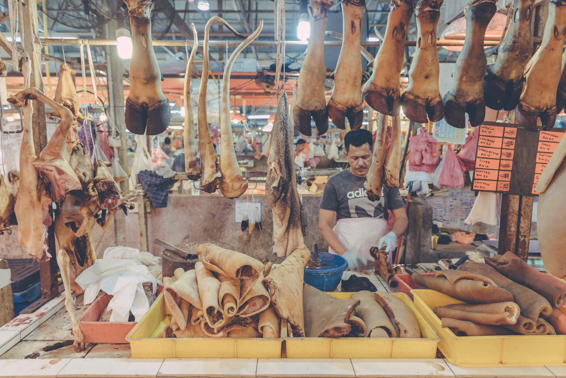 Butcher seated behind a Malaysian market stall displaying hanging meat and animal parts.