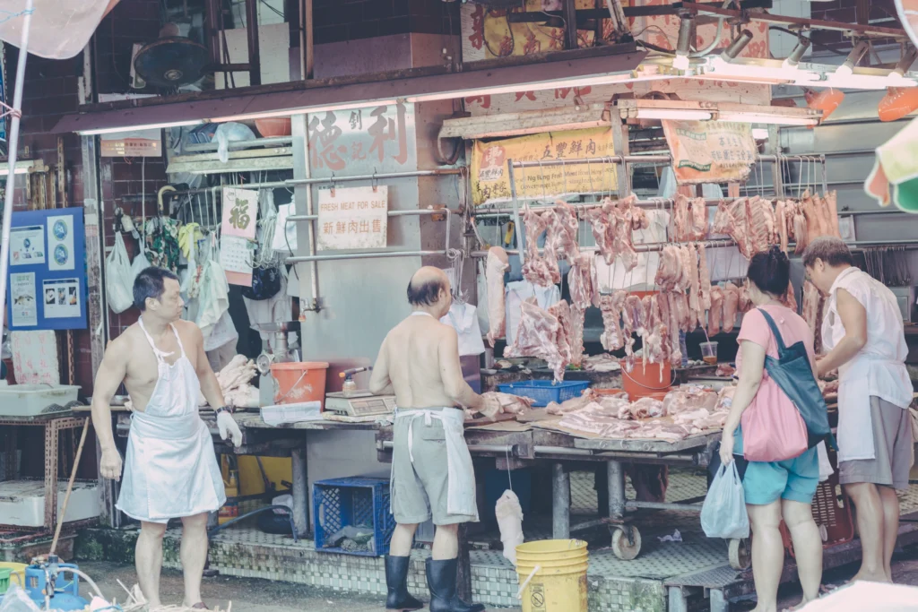 Customers and vendors at an open butcher stall in a Hong Kong wet market, with cuts of meat hanging above the counter.