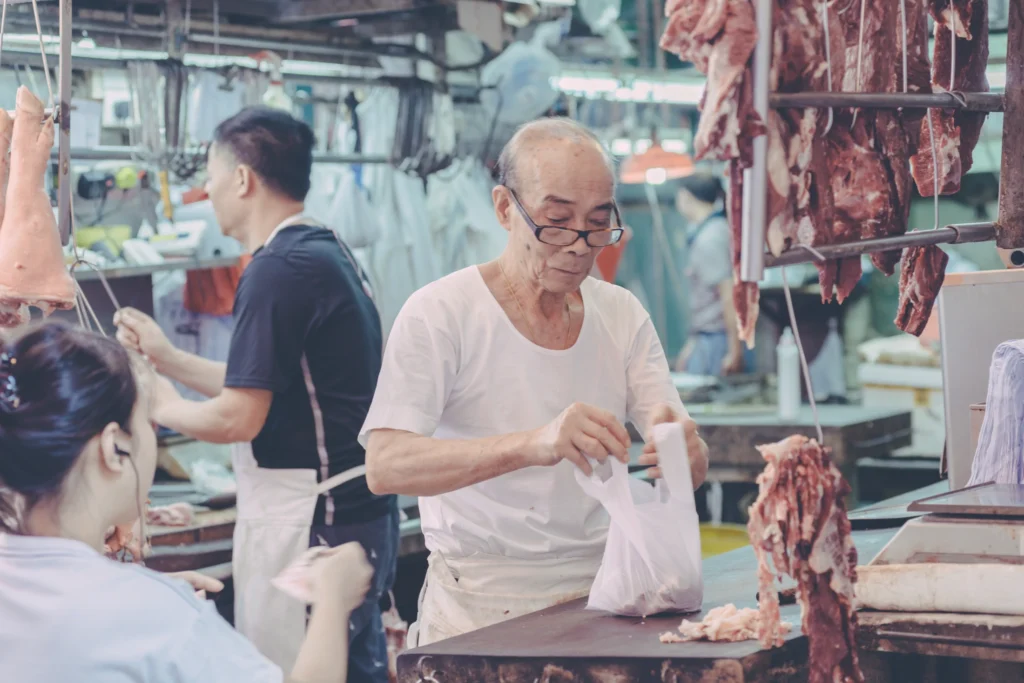 Vendor handing a purchase to a customer at a Hong Kong meat stall, surrounded by hanging cuts of meat.