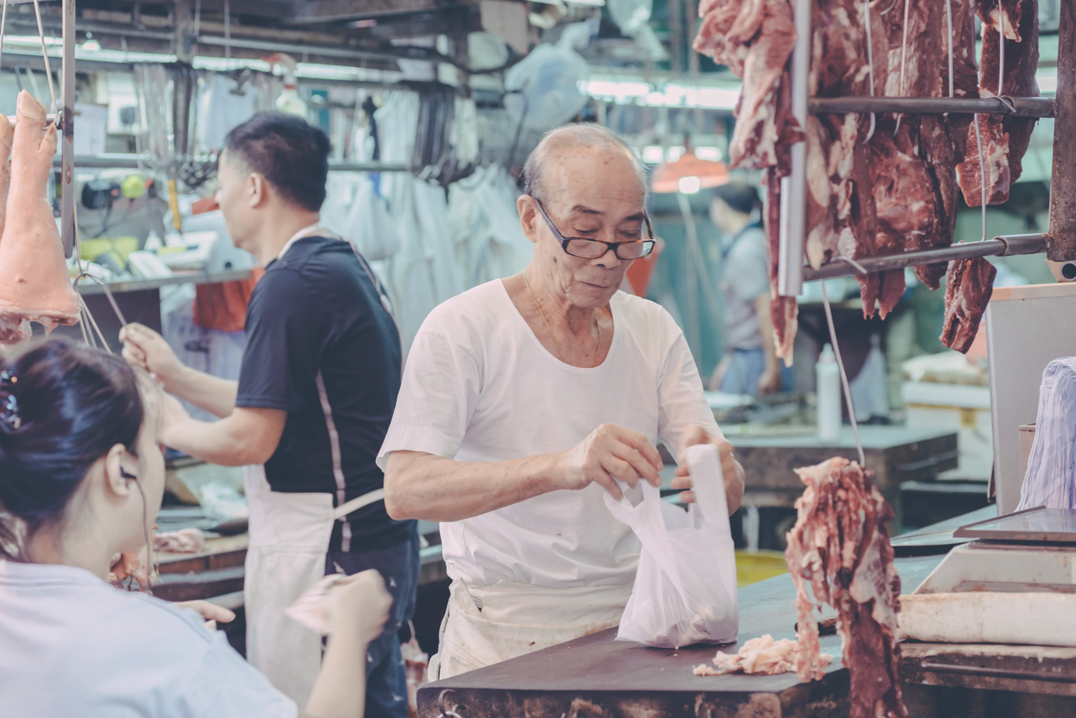 Vendor handing a purchase to a customer at a Hong Kong meat stall, surrounded by hanging cuts of meat.