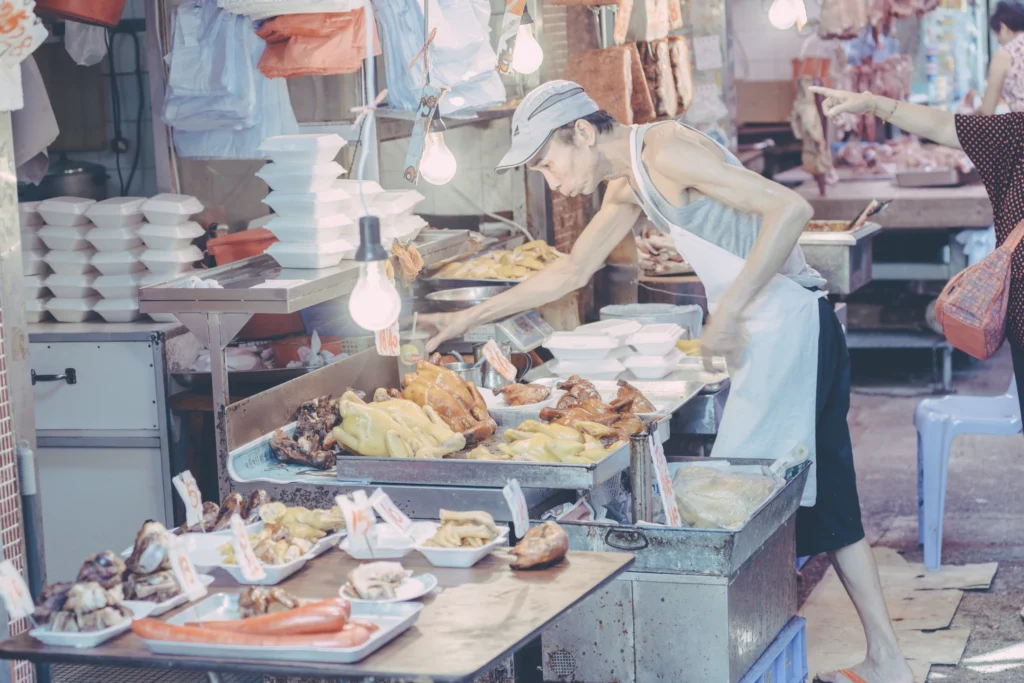 Market vendor reaching across a counter filled with prepared food and fresh produce in a Hong Kong wet market.