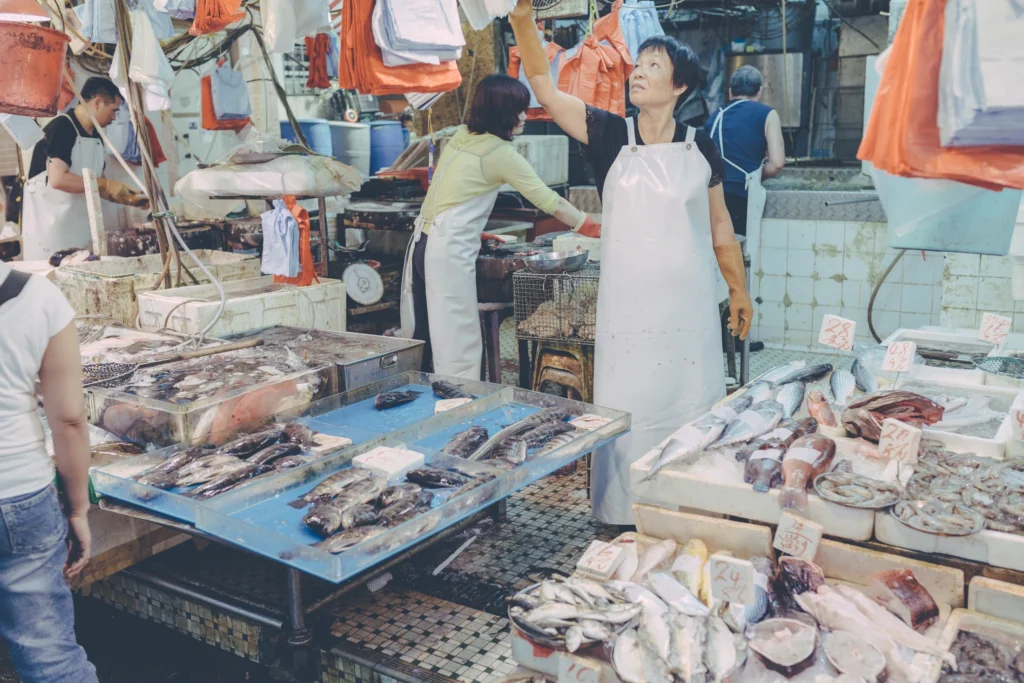 Seafood vendor holding an orange bag above a crowded fish stall in a Hong Kong wet market.