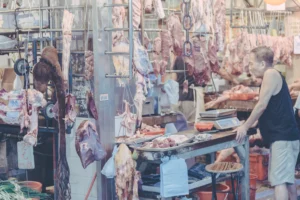 Butcher working behind a market counter framed by hanging cuts of meat in Hong Kong.