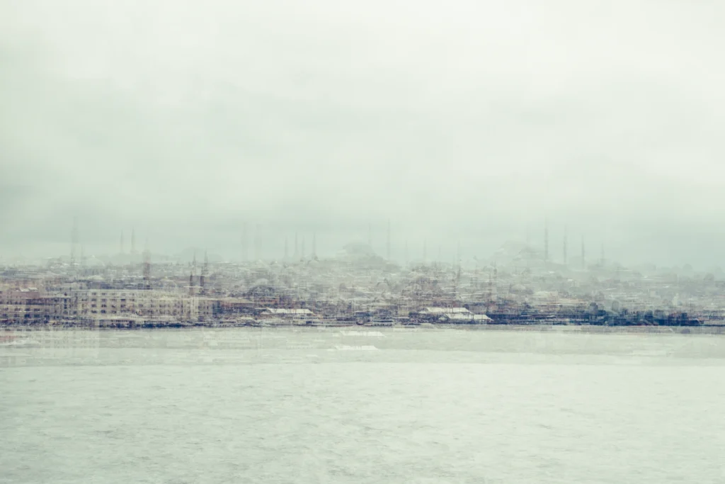 Pale multilayer photograph of Istanbul’s waterfront across the water, with overlapping historic buildings and the Galata Tower emerging through a hazy skyline.