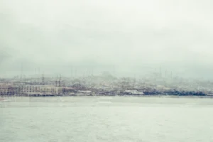 Pale multilayer photograph of Istanbul’s waterfront across the water, with overlapping historic buildings and the Galata Tower emerging through a hazy skyline.