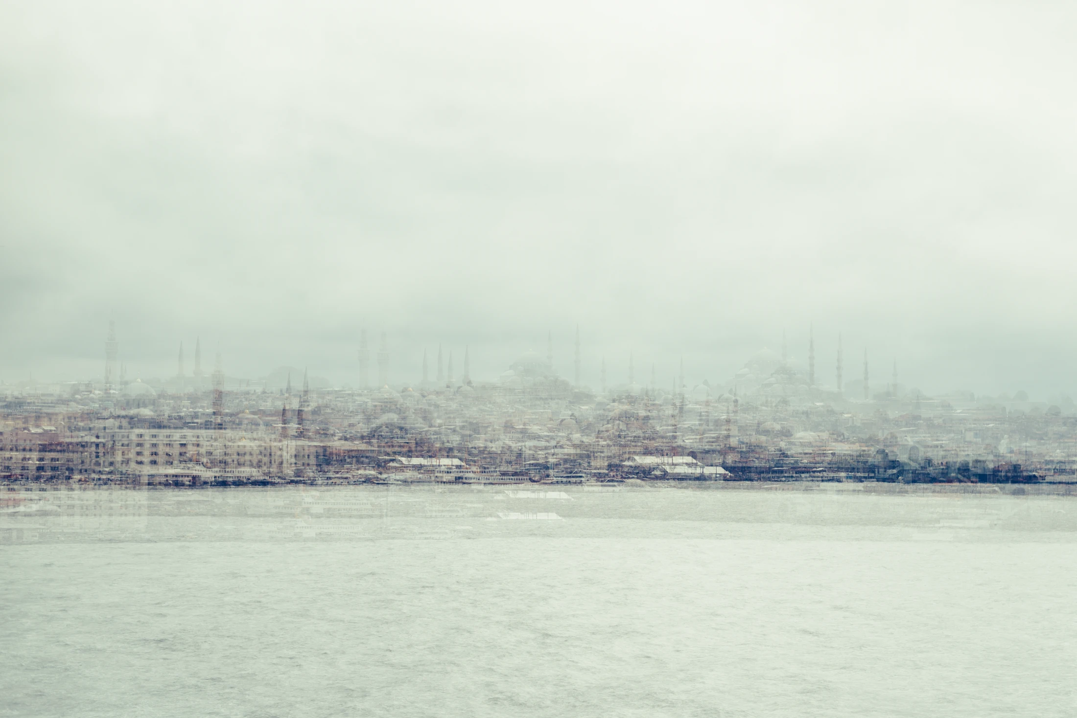 Pale multilayer photograph of Istanbul’s waterfront across the water, with overlapping historic buildings and the Galata Tower emerging through a hazy skyline.